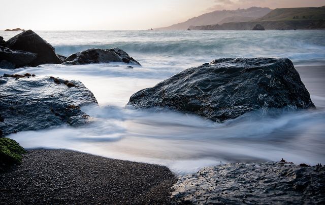 Surf swirls around dark boulders on Goat Rock Beach as the evening light fades